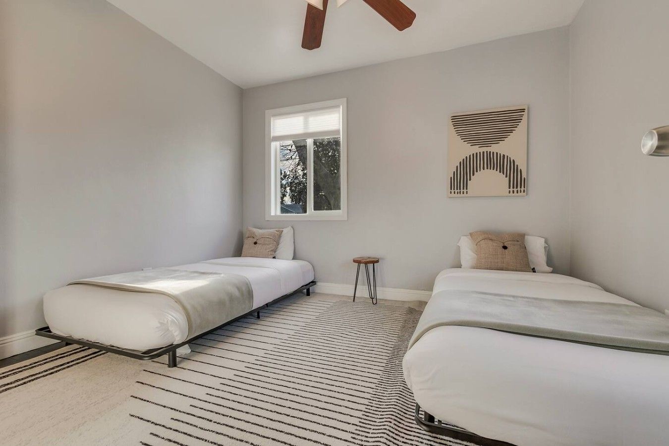 A bedroom with two beds, a window, a small stool, and a ceiling fan.
