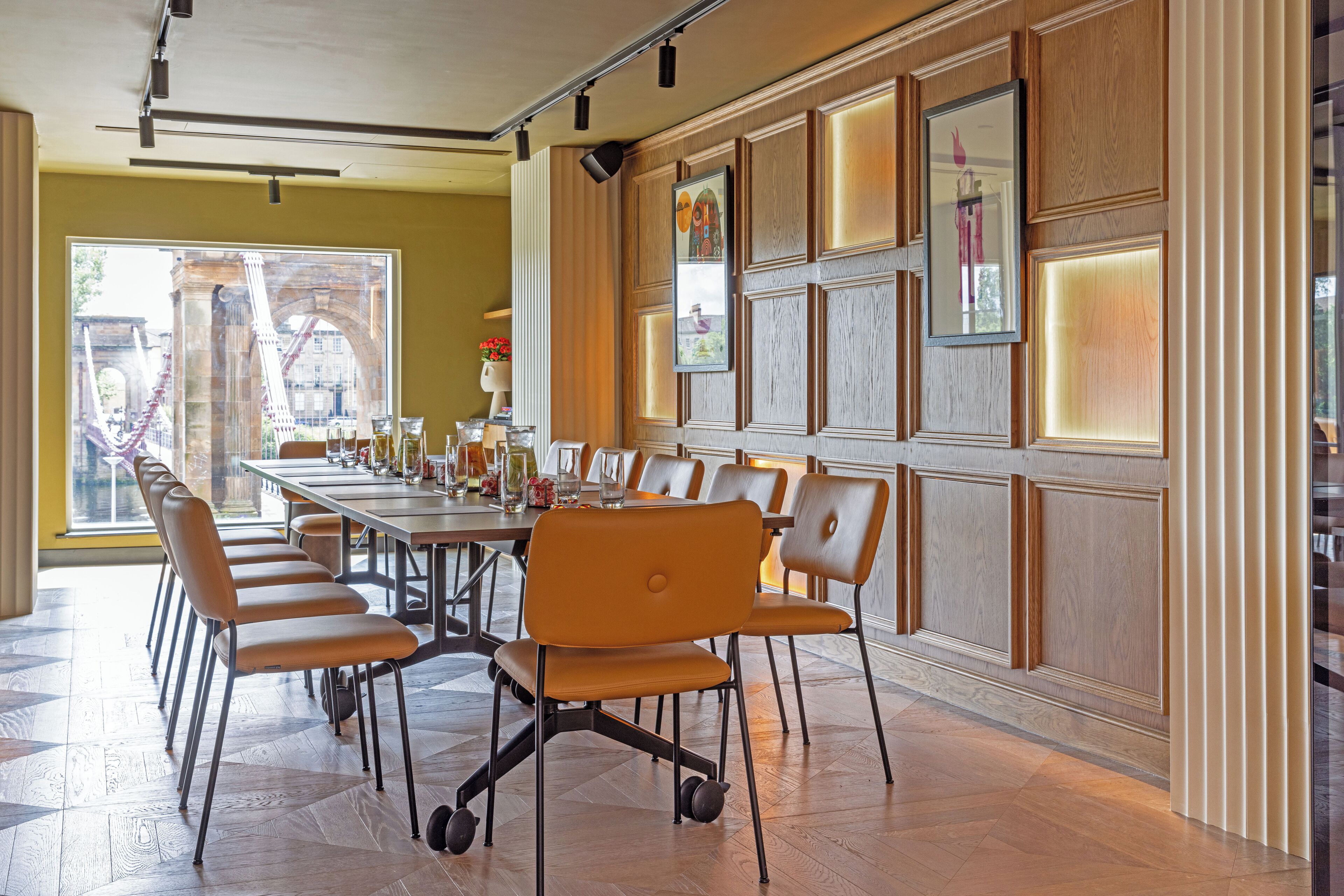 A conference room with a long table and multiple chairs, large windows with a view of a bridge, and wooden paneling on the walls.