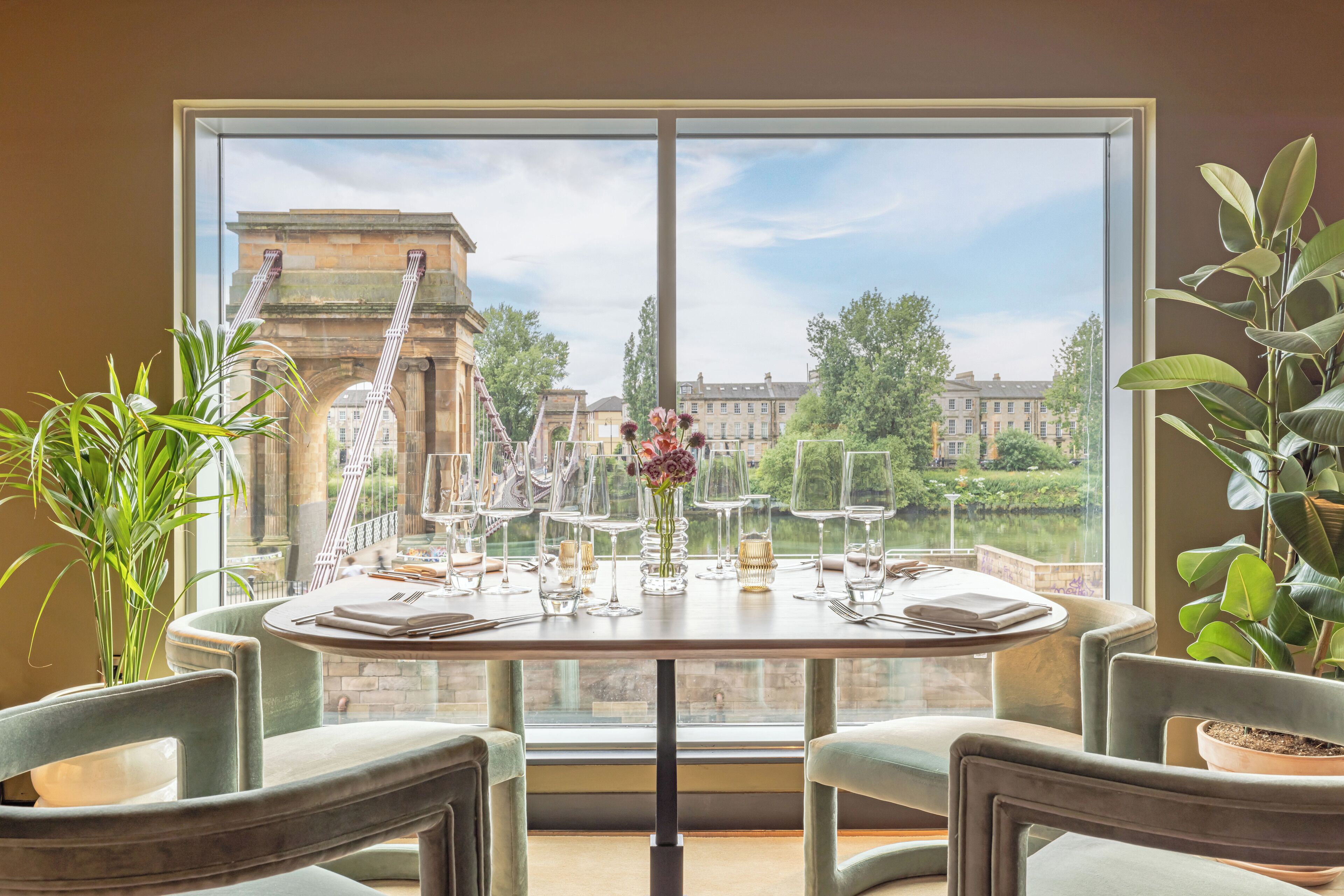 Mesa de comedor dispuesta con vasos y cubiertos, rodeada de sillas, con vistas a un edificio histórico y vegetación a través de amplios ventanales.