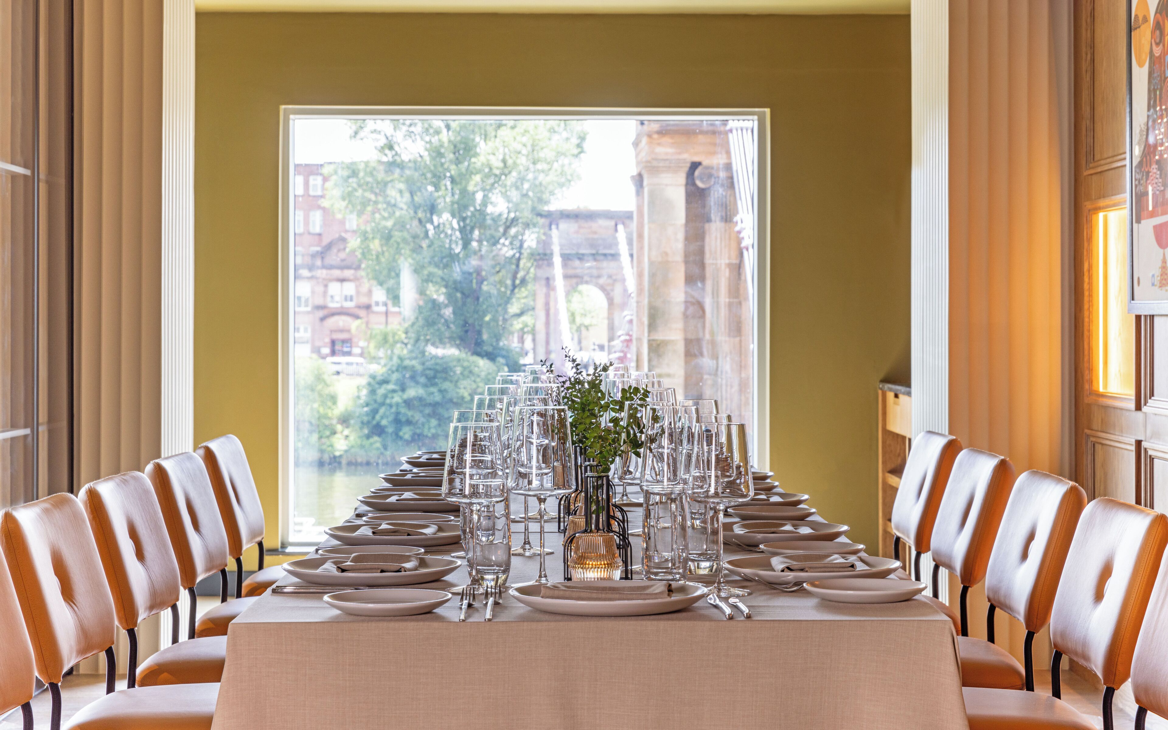 A dining table set for a meal with chairs on either side, a vase with greenery, and a view of a building through a window.