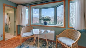 Une pièce avec une table en verre, deux chaises en osier, un radiateur et une vue sur l’extérieur grâce à de grandes fenêtres.