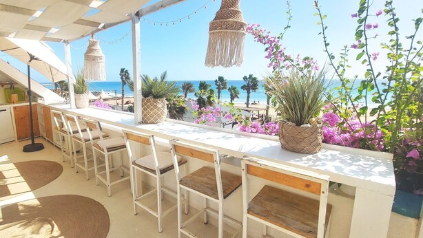 Zona d'exterior per dinar a plena llum del sol, amb una taula i cadires blanques, amb vista a una platja.