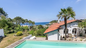 A white villa with a red-tiled roof overlooking a pool and a coastal view.