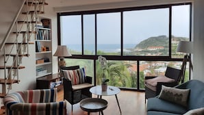 A spacious living room with a large window offering a view of the sea and a hilly landscape.