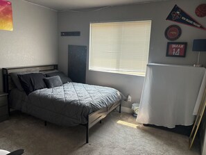 A bedroom with a bed, a window with blinds, a wall-mounted shelf, and a framed sports jersey.