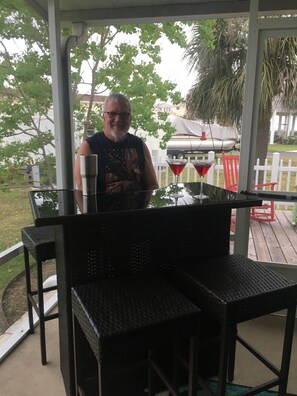 Ein überdachter Außenbereich mit Tisch und Stühlen, einem Grill und einem weißen Zaun im Hintergrund.