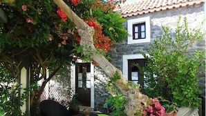 A stone building with a tiled roof, a wooden door, and a window. There is a tree with red flowers and a chair with green cushions. - Cottage/Gite Cavalinho (Calheta)