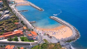 An aerial view of a coastal area with a beach, a curved breakwater, and buildings with red roofs. - Cottage/Gite Cavalinho (Calheta)
