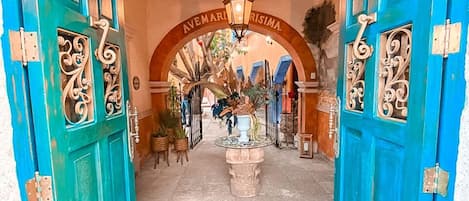 Un arco turquesa que da a un patio con una fuente, rodeado de plantas en macetas y un techo de madera.