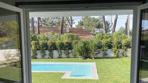 Vista a través de un ventanal corredero hacia un jardín trasero con piscina, césped y una casa al fondo.