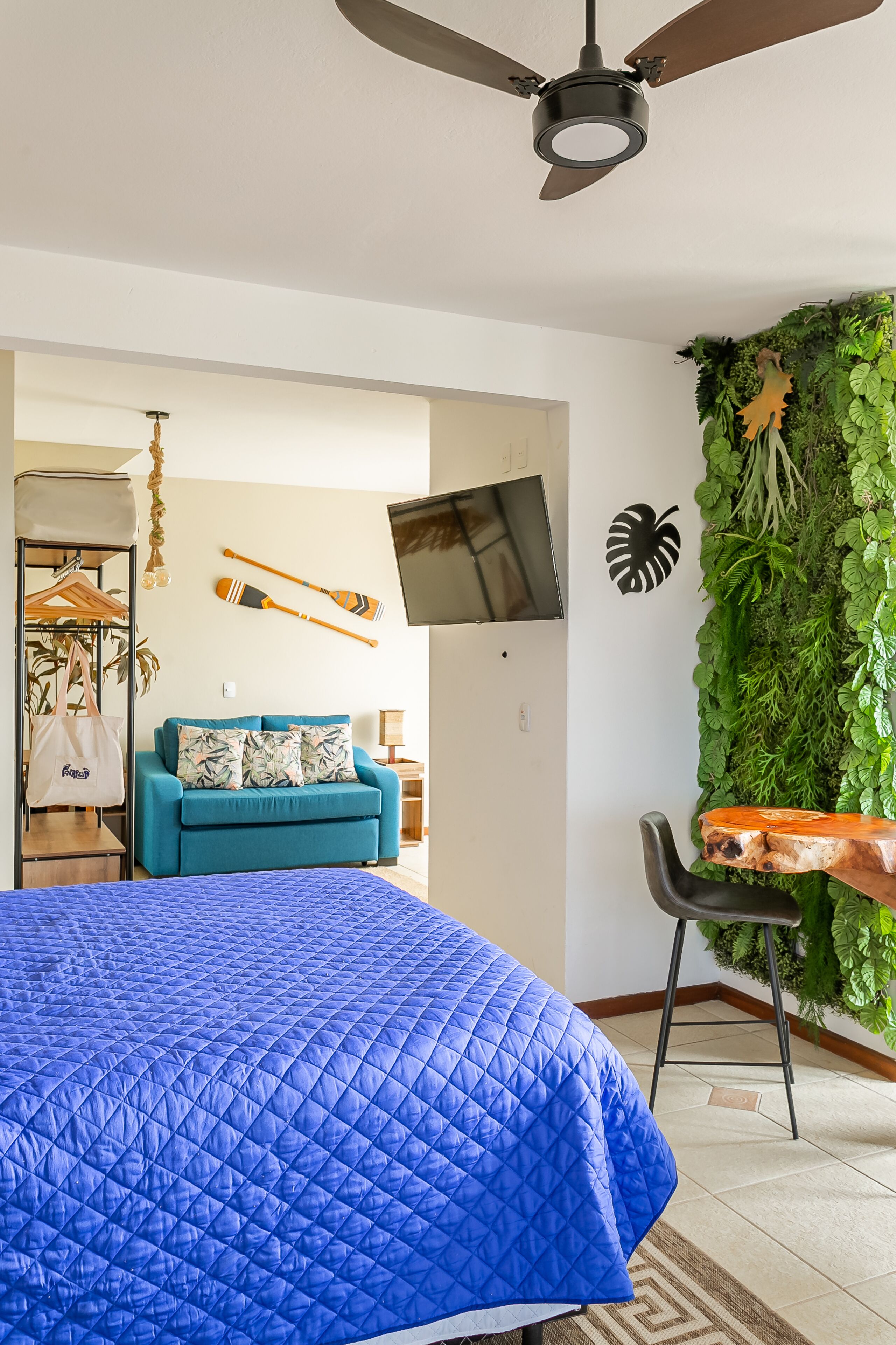 A modern bedroom with a blue quilted bedspread, a teal sofa with tropical pillows, a wooden side table, a mounted TV, and a green vertical garden wall.
