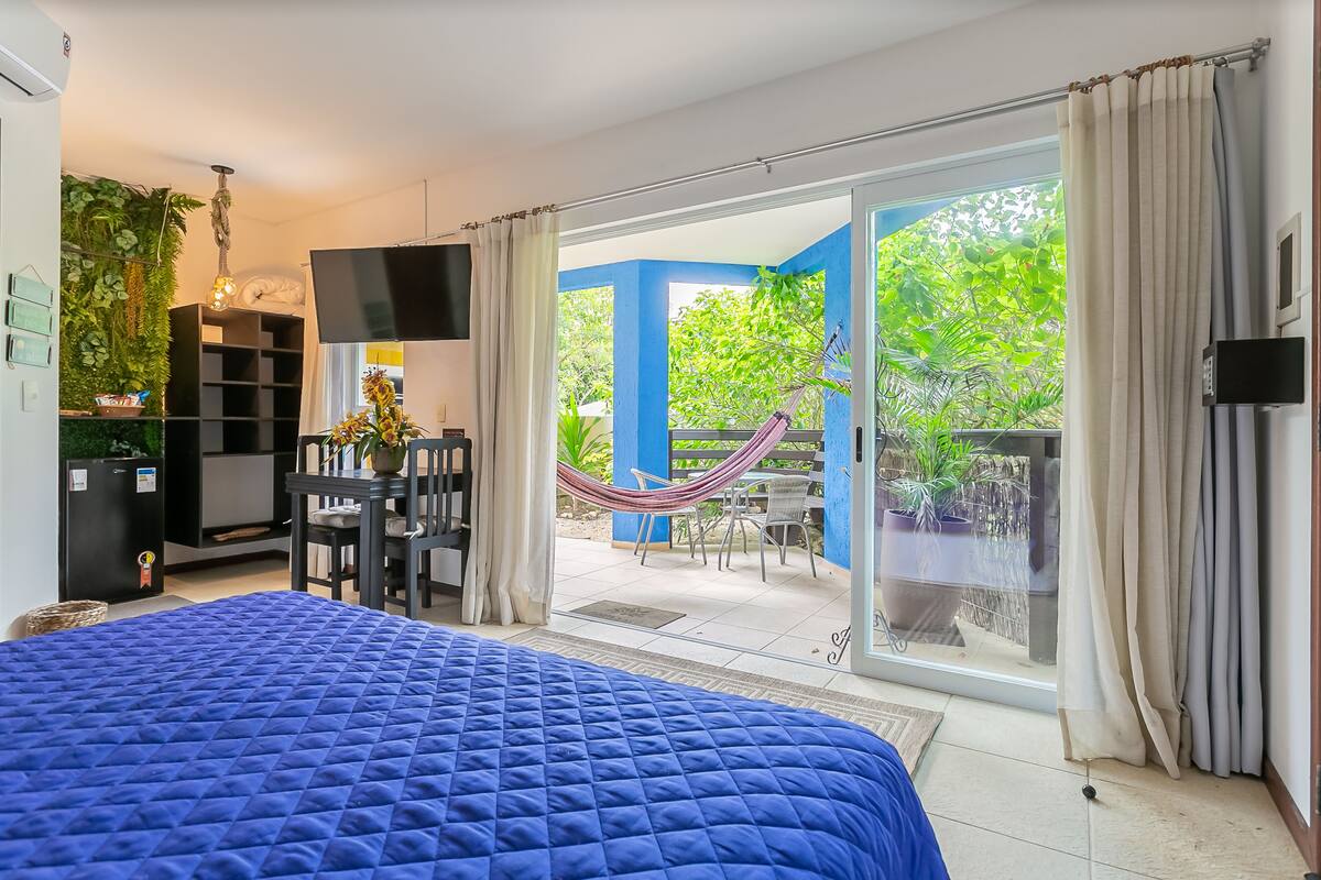 A bedroom with a large bed, a TV, a desk, and a view of a patio with a hammock and plants.