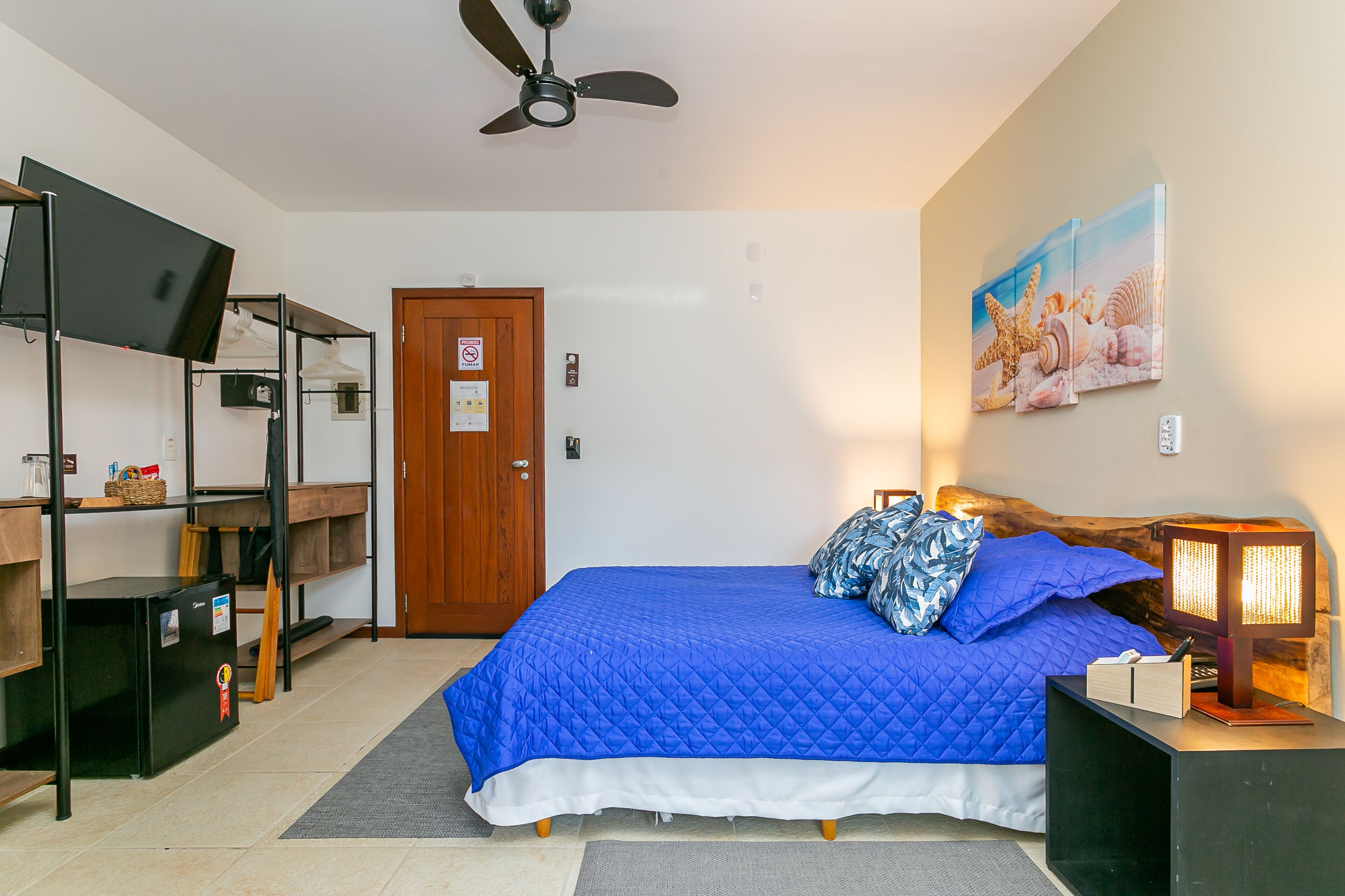 A bedroom with a bed, a ceiling fan, a wooden door, a wall-mounted shelf, and a nightstand with a lamp.
