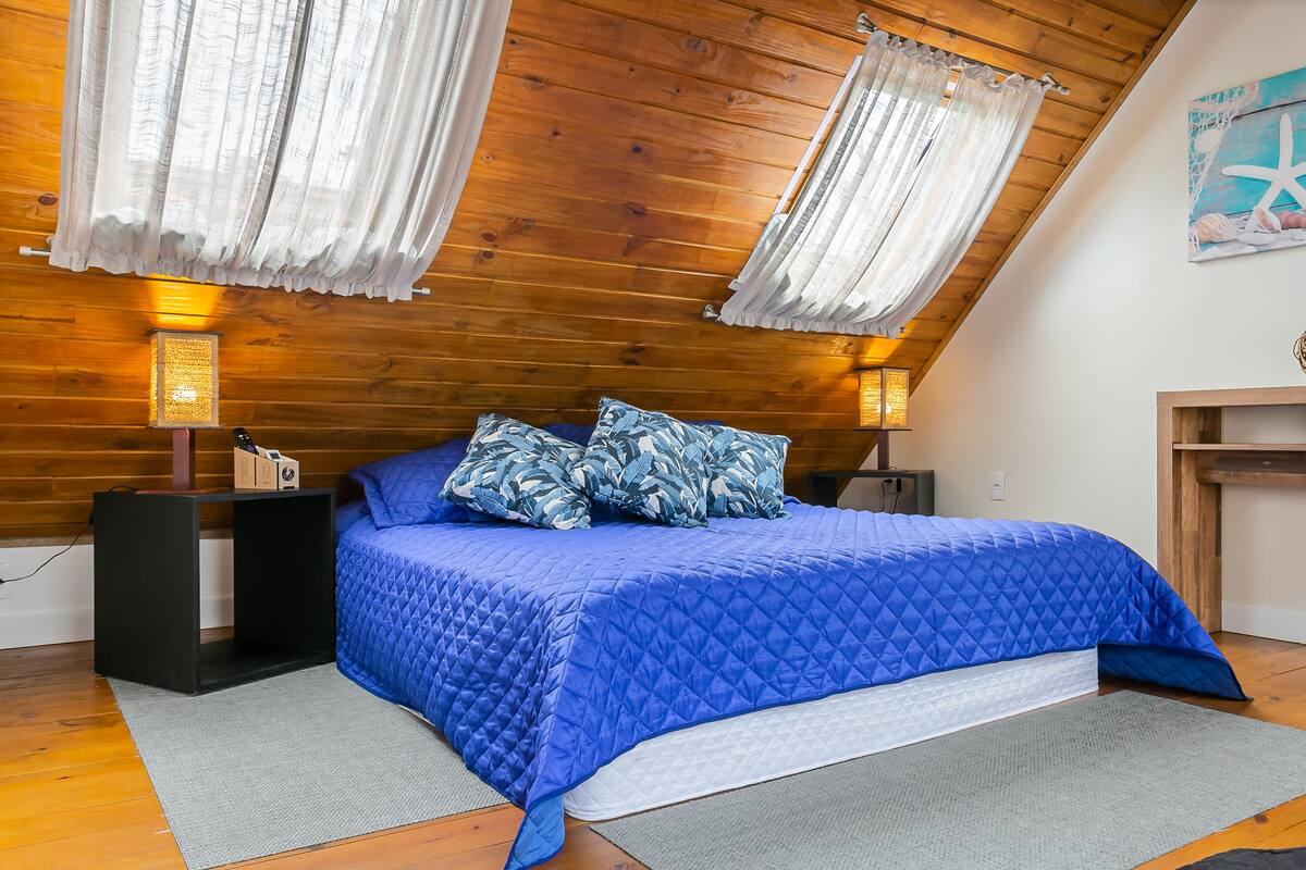 A bedroom with a wooden ceiling, a blue quilted bedspread, and a nightstand with a lamp.