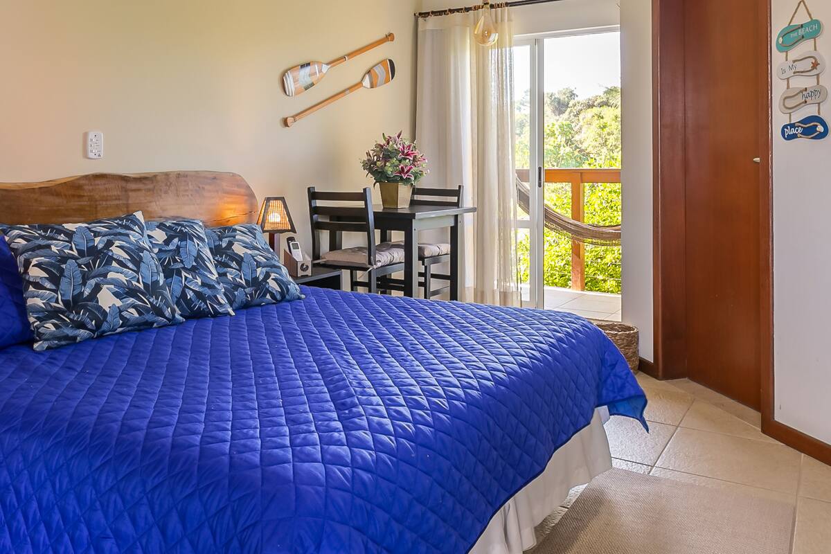 A bedroom with a large bed, a wooden headboard, a table with chairs, a hammock, and a view of greenery outside.