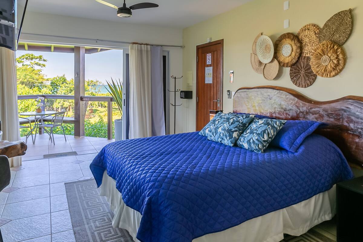 A bedroom with a large bed, a ceiling fan, a view of the outdoors, a wooden door, and a wall with woven decorations.