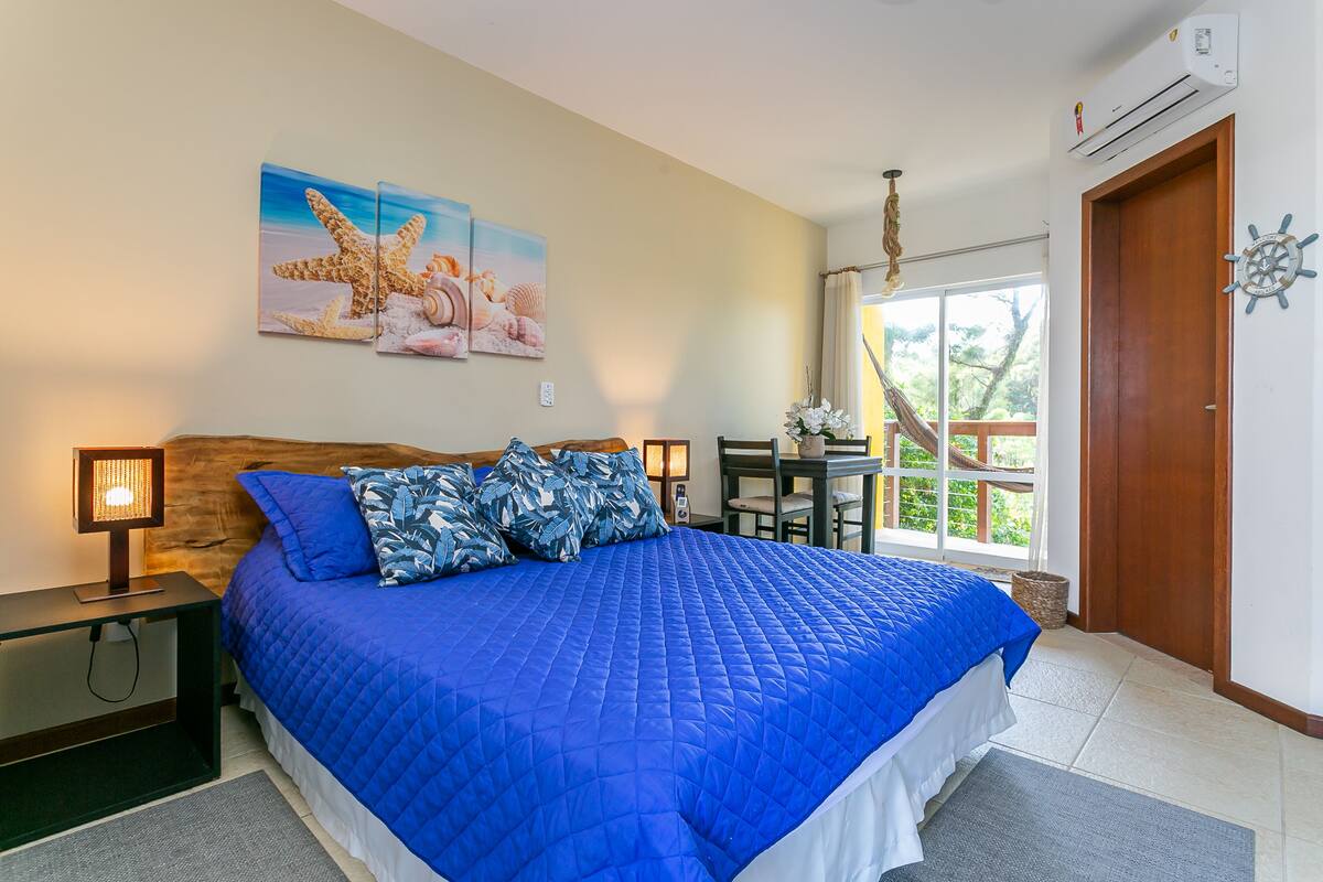 A bedroom with a large bed, a wooden headboard, a nightstand with a lamp, a window with a view of greenery, and a door with a decorative item.