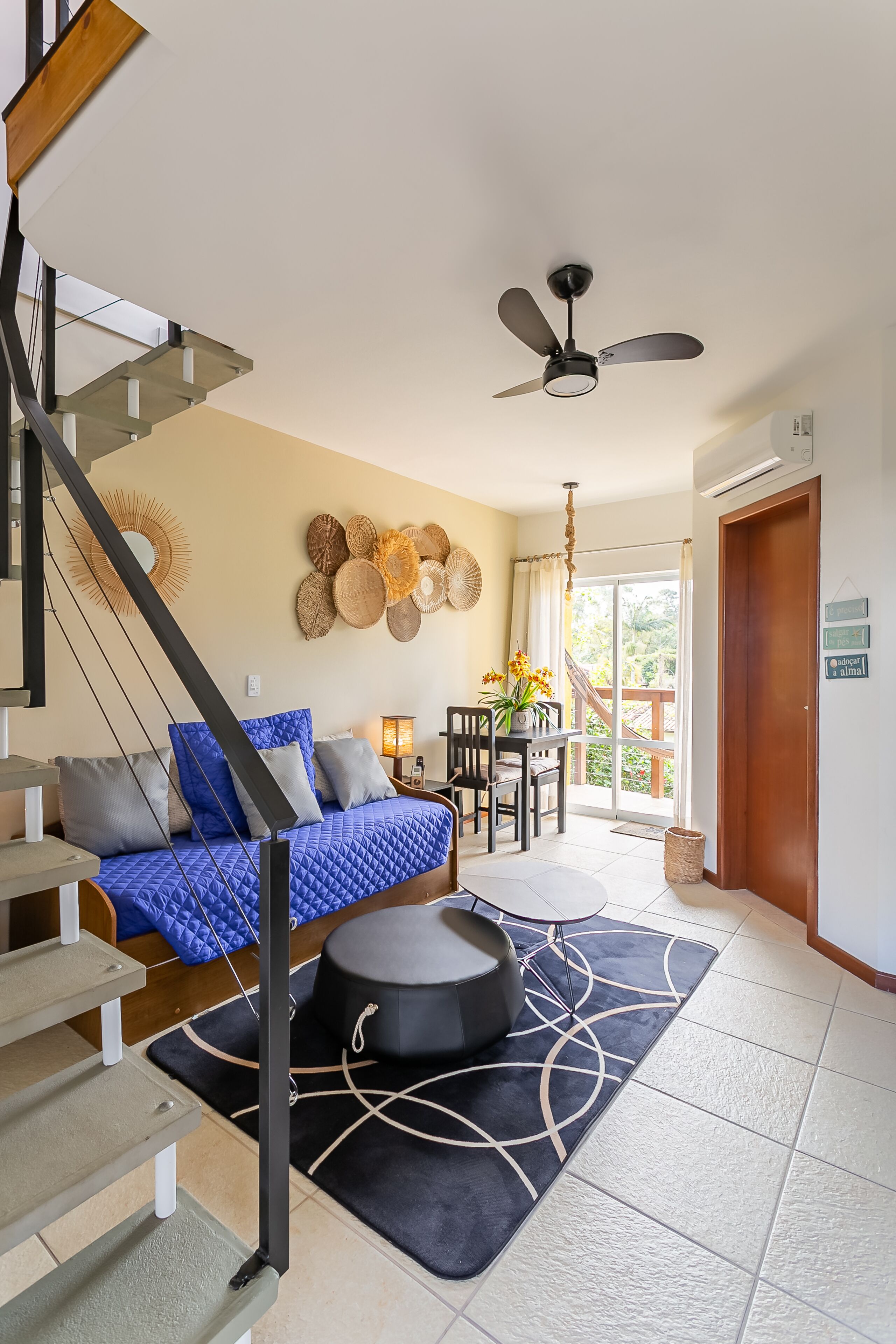 Une chambre moderne avec un escalier, un lit avec une literie bleue et grise, un coin repas avec une table et des chaises, et un ventilateur de plafond.