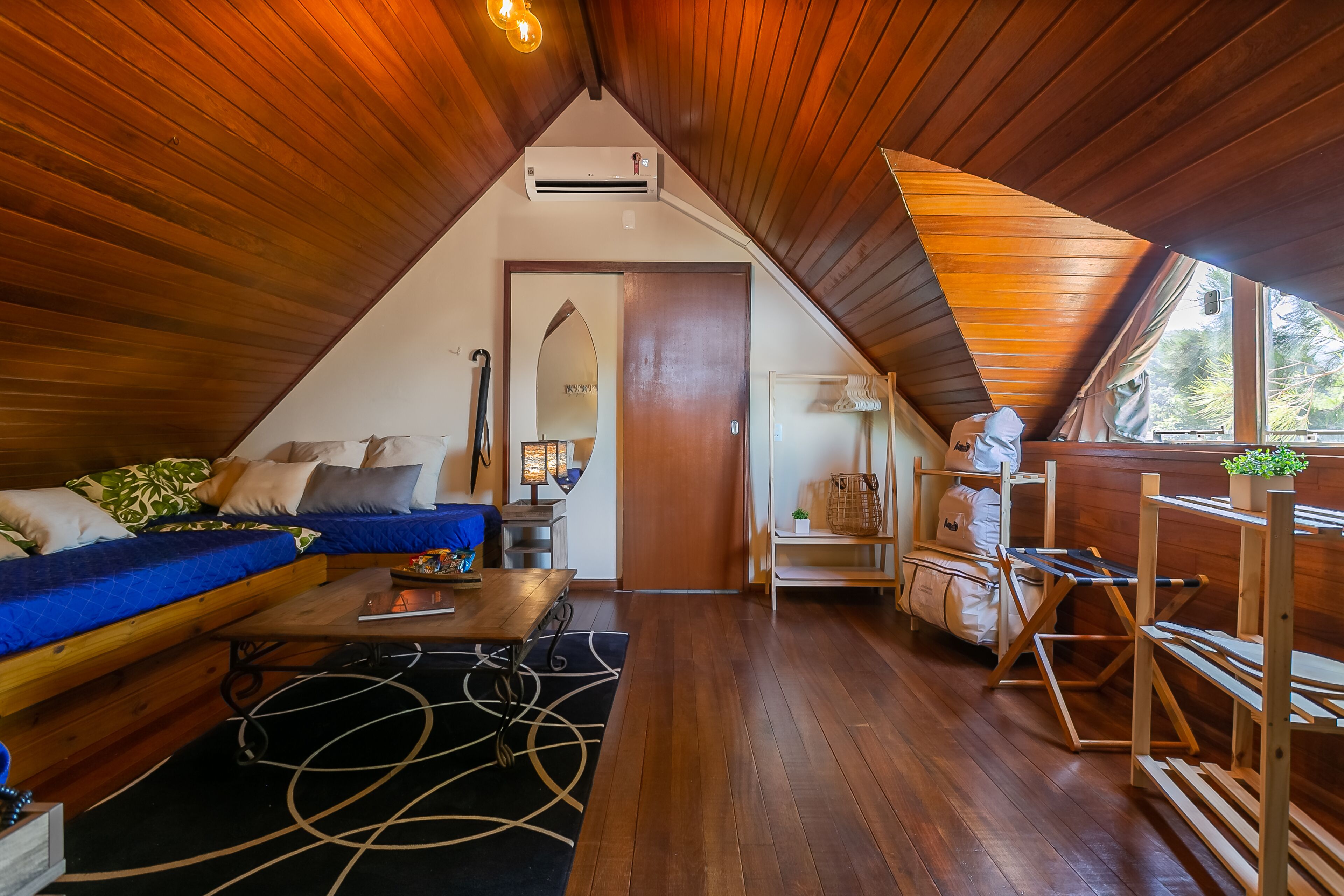 A cozy attic room with a wooden ceiling, a sofa with patterned cushions, a small table, and a window with a view of trees.