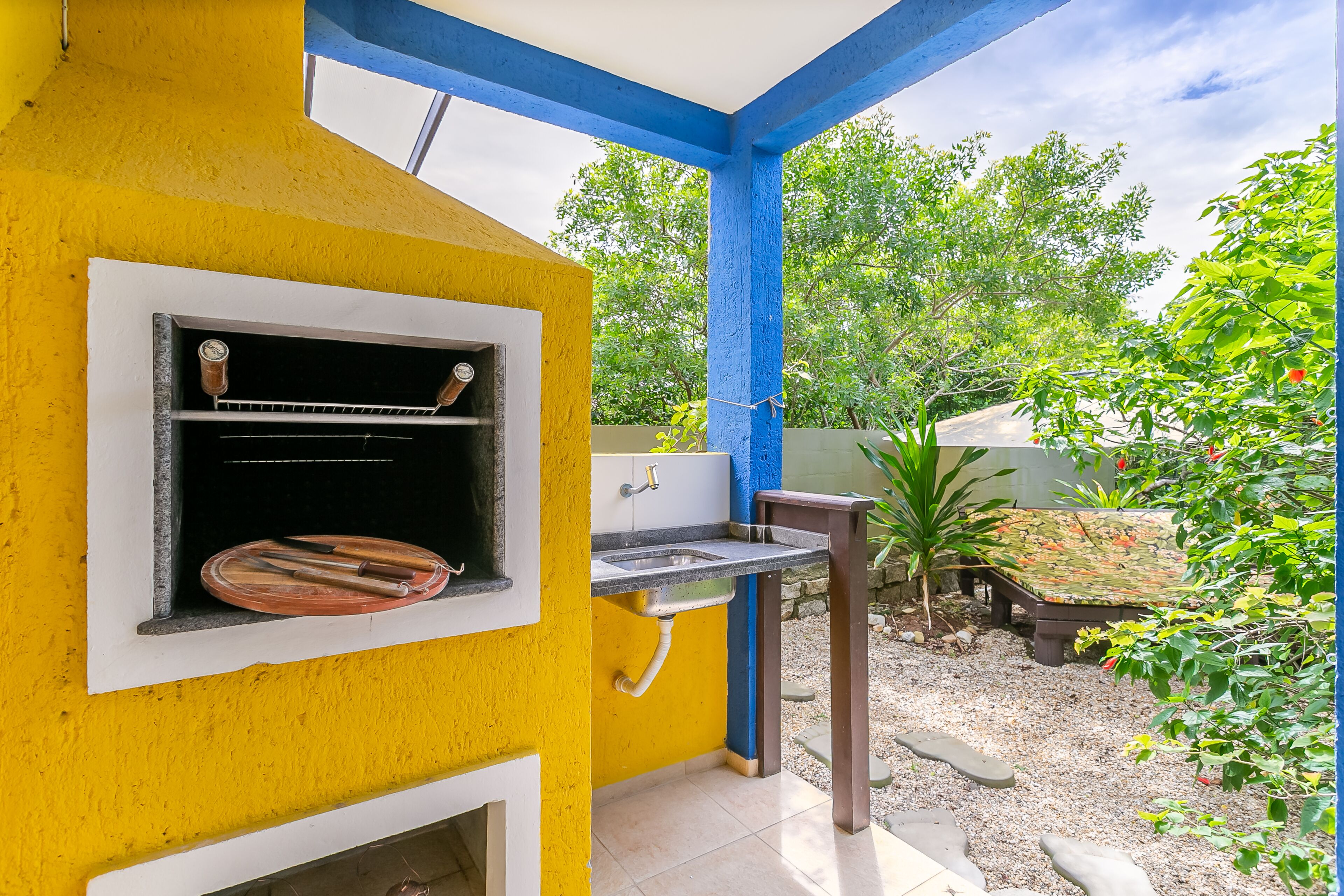 A small outdoor kitchen with a grill, a sink, and a window with a plate of utensils.
