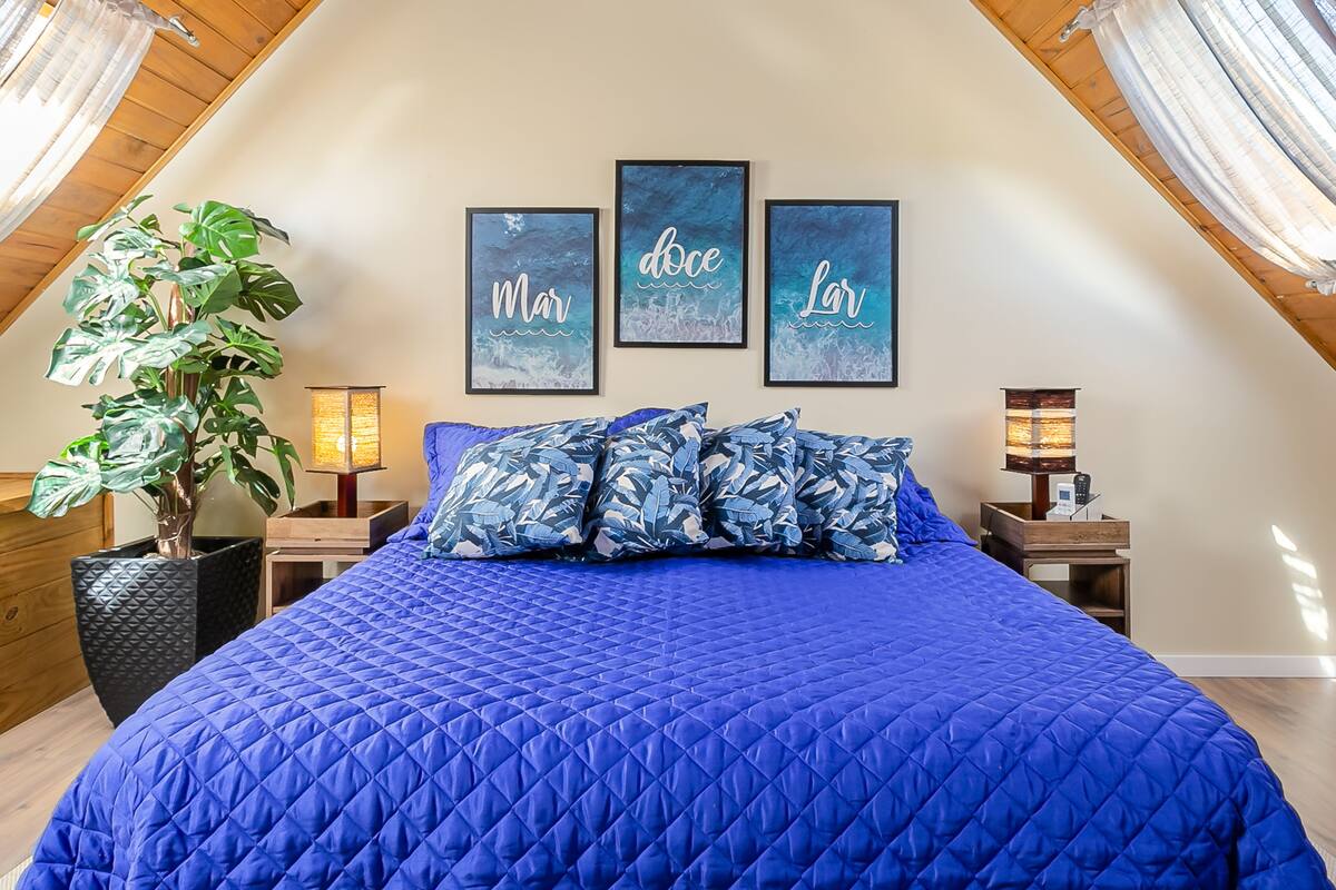 A bedroom with a sloped ceiling, a large bed with blue bedding, a potted plant, and three framed artworks on the wall.