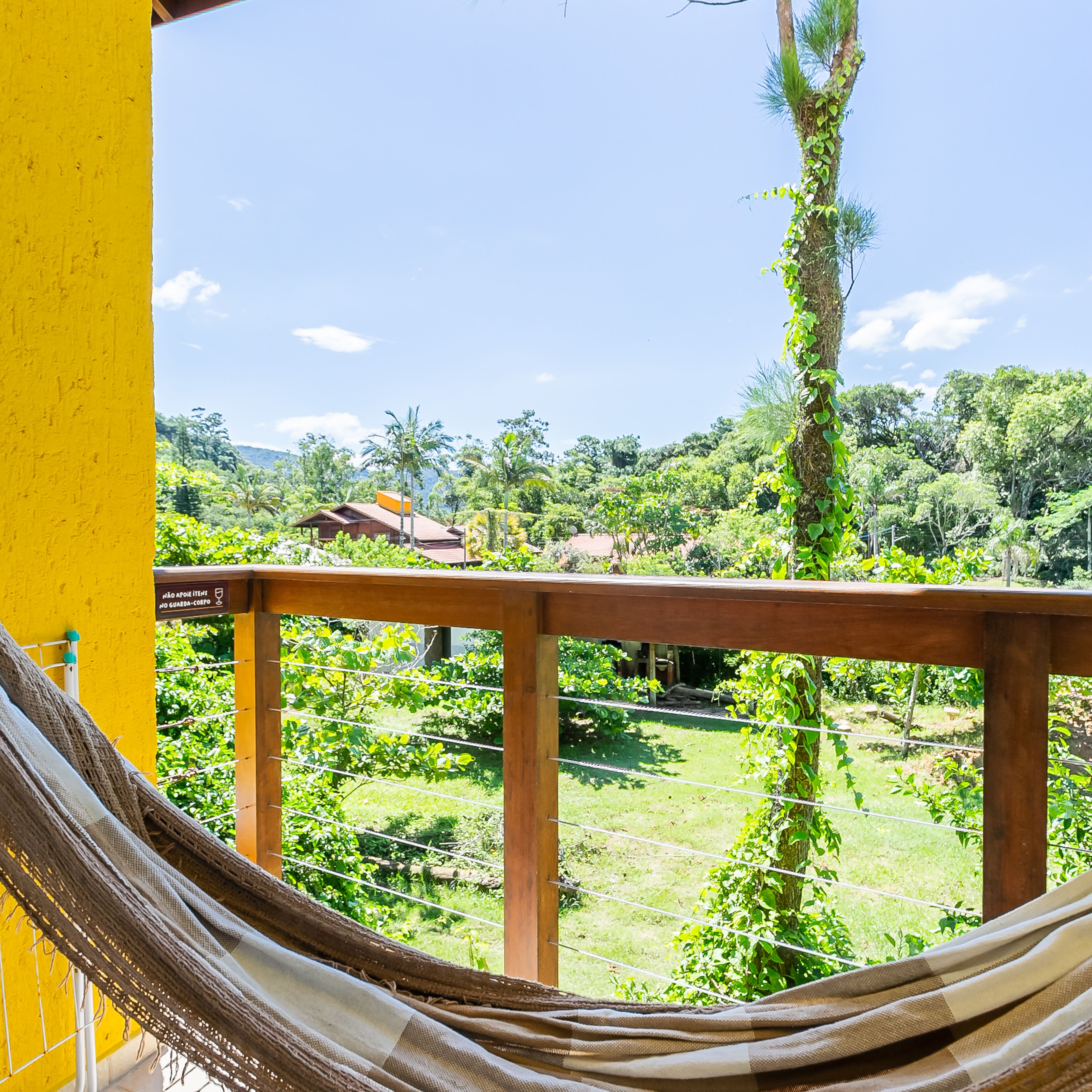 Un hamac sur un balcon donnant sur un jardin luxuriant, avec une balustrade en bois et un mur jaune.