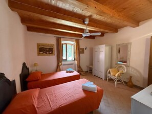 A room with two beds, wooden ceiling beams, a ceiling fan, a window with curtains, a white wardrobe, a chair, and a mirror.