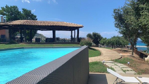 A swimming pool with a pergola and outdoor seating area, surrounded by greenery and a view of the sea.