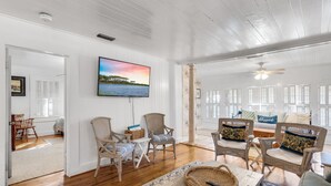 Een ruime woonkamer met witte wandpanelen, een grote flatscreen televisie boven een rieten fauteuil, en een plafondventilator.