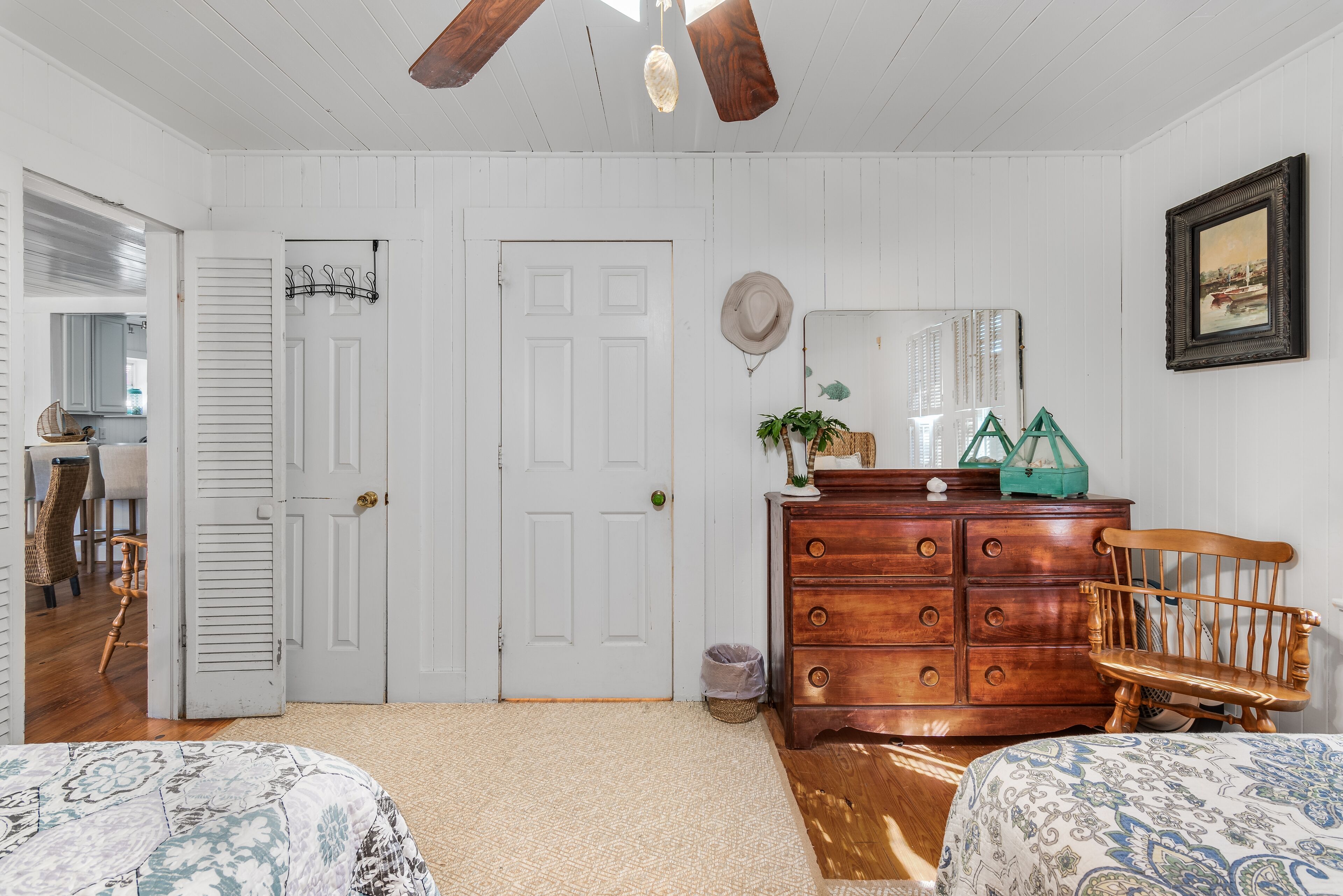 A bedroom with a wooden dresser, two beds, a ceiling fan, and a door leading to another room.