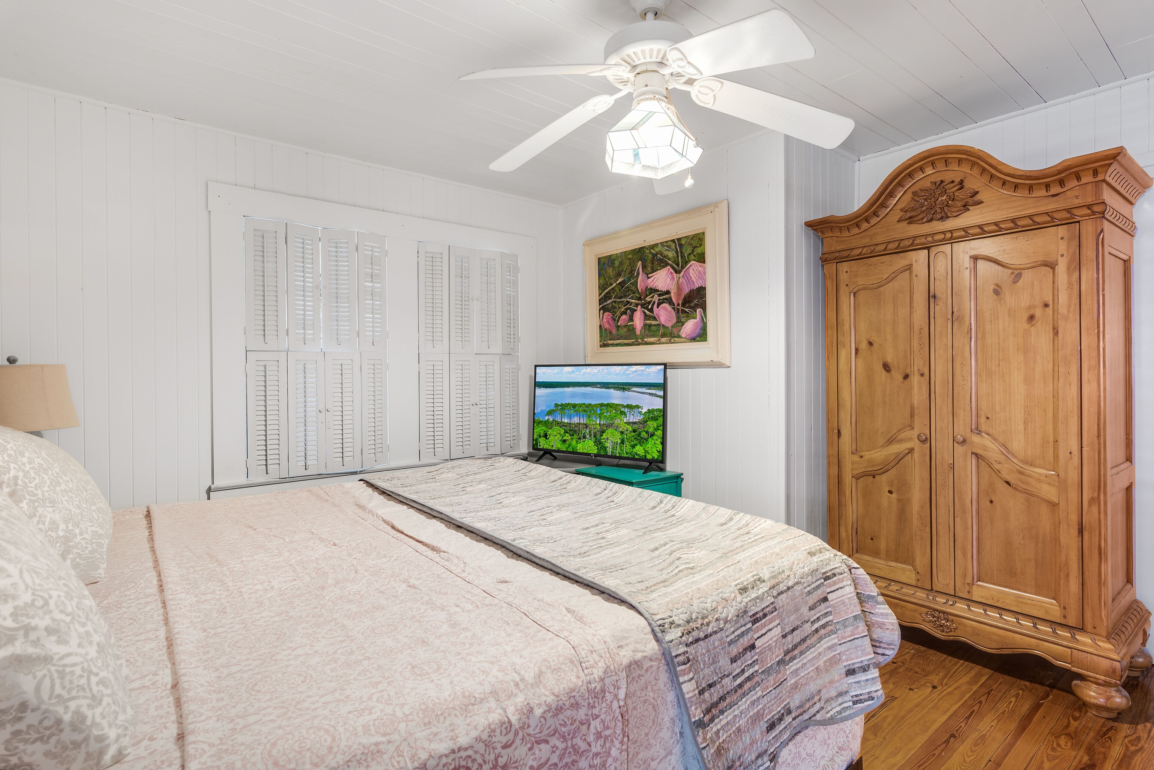 Une chambre à coucher avec un lit, une armoire en bois, un ventilateur de plafond, un tableau encadré et un téléviseur posé sur un meuble.