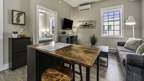 A modern living room with a wooden dining table, a grey sofa, and a flat-screen TV mounted on the wall. - Cozy Efficiency at Downtown Manistee (Manistee)