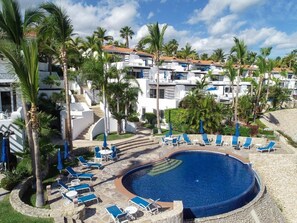 Zona de piscina con sillones de descanso y sombrillas, rodeada de palmeras y un edificio con varios balcones.