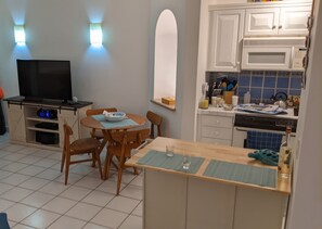 A compact kitchen with white cabinets, a dining area with wooden chairs, a TV, and a small island with a glass.