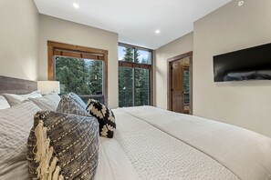 A modern bedroom with a large bed, a TV mounted on the wall, and large windows offering a view of trees.