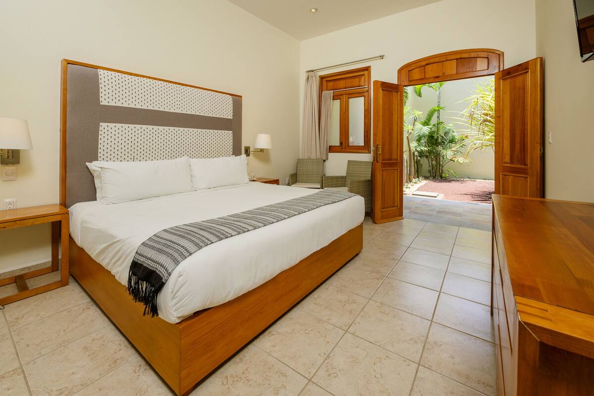 Une chambre à coucher bien aménagée, avec un grand lit, des tables de chevet, une armoire en bois et une vue sur un jardin par une porte ouverte.