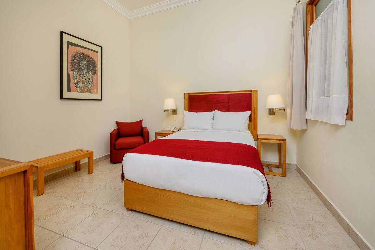 Une chambre d’hôtel bien rangée, avec un grand lit, un fauteuil rouge, deux tables de chevet avec des lampes et une œuvre d’art encadrée accrochée au mur.