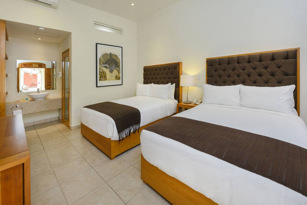 Une chambre d’hôtel avec deux lits, une salle de bain avec un lavabo et un miroir, et un tableau encadré au mur.
