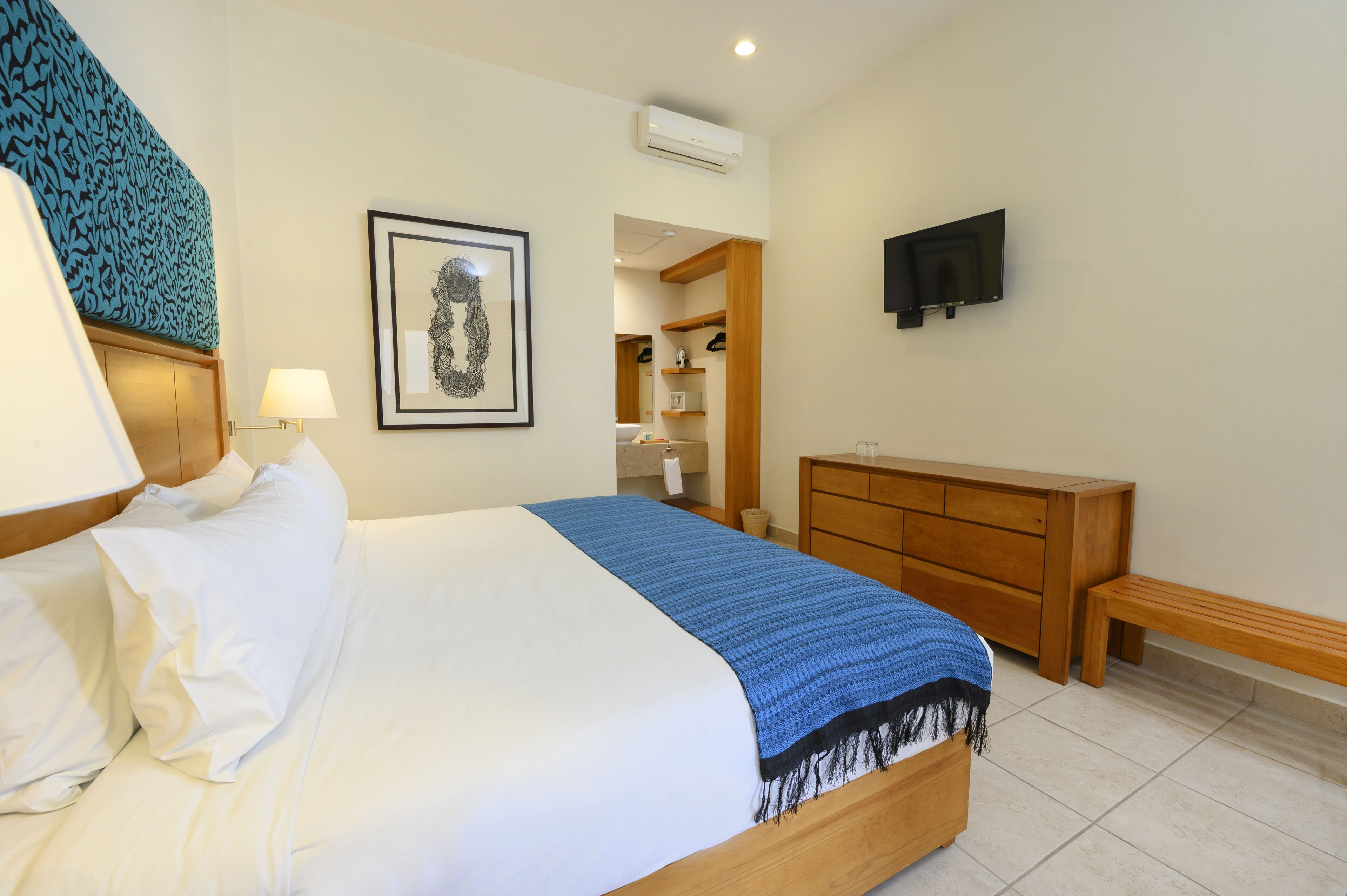 Una habitación de hotel con una cama, un aparador de madera, un televisor montado en la pared y una obra de arte enmarcada.