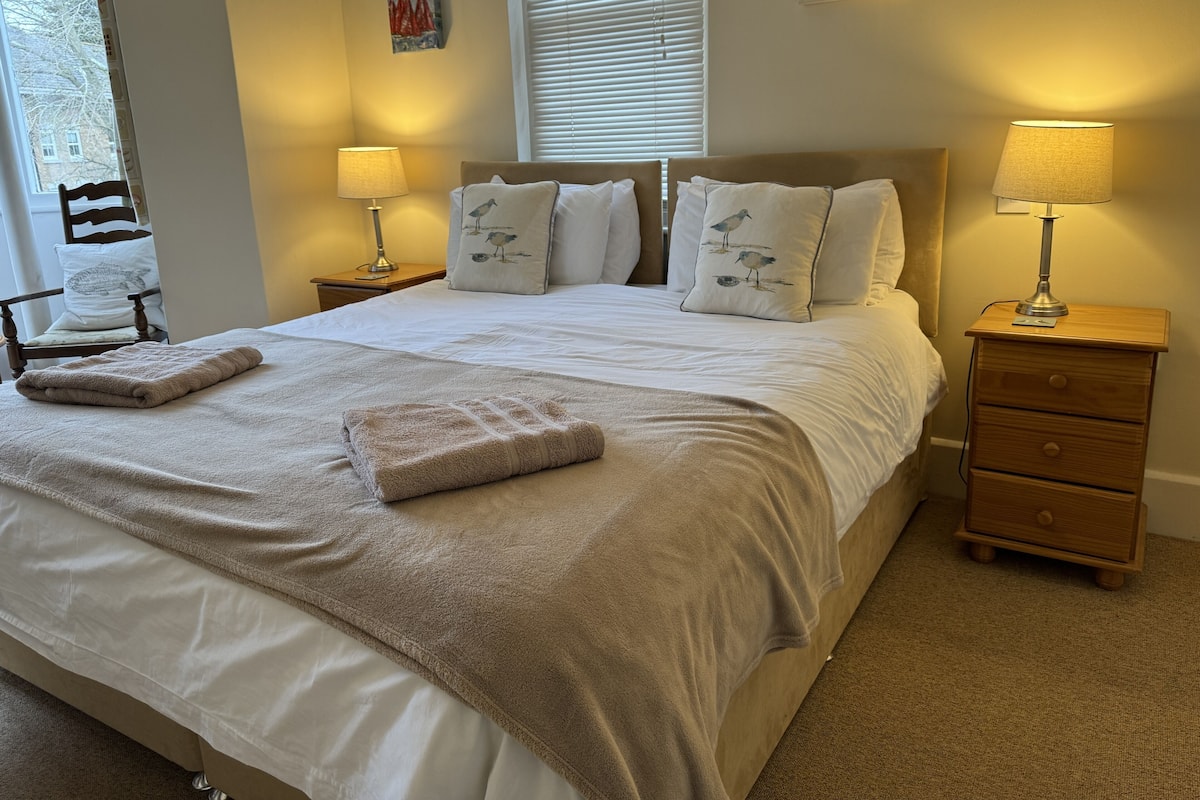 Una cama bien hecha con una manta color beige y almohadas, dos lámparas de noche, una mesita de madera y una silla con cojín.