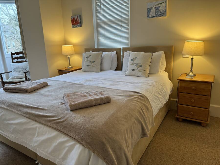 A neatly made bed with a beige blanket and pillows, two bedside lamps, a wooden nightstand, and a chair with a cushion.
