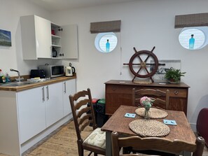 A compact kitchen with white cabinets, a wooden dining table with woven placemats, and a ship's wheel decoration. - The Pilot Boat Inn (Bembridge)
