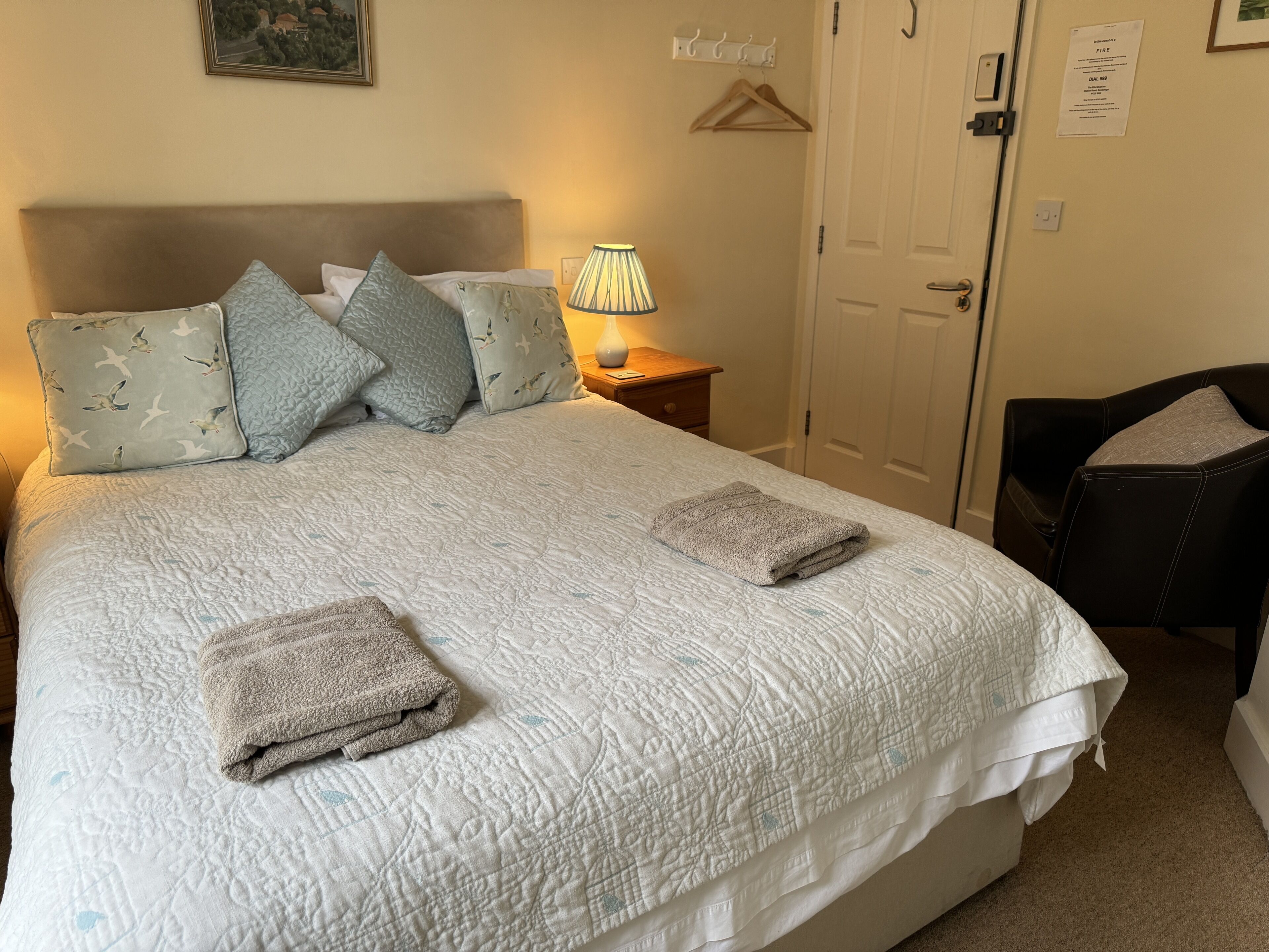 Un lit bien fait, avec une couette blanche matelassée, plusieurs oreillers et deux serviettes pliées. On y trouve une lampe de chevet sur une table de nuit en bois, une porte sur laquelle est apposée une notice de sécurité incendie, et une chaise avec un coussin.