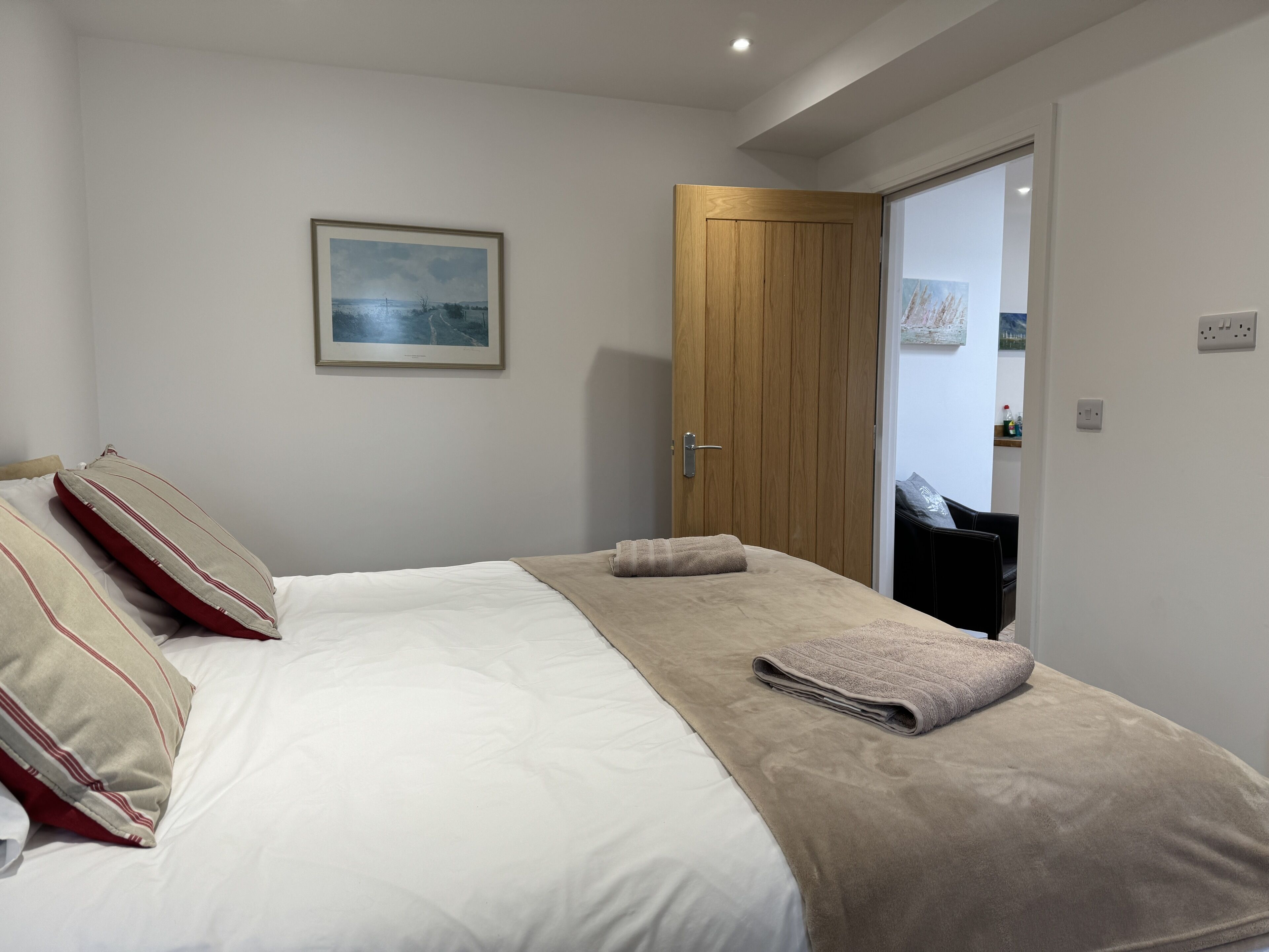 A neatly made bed with a white comforter and two pillows, a beige blanket, and a folded towel. A wooden door is visible to the right, and a framed picture hangs on the wall above the bed.