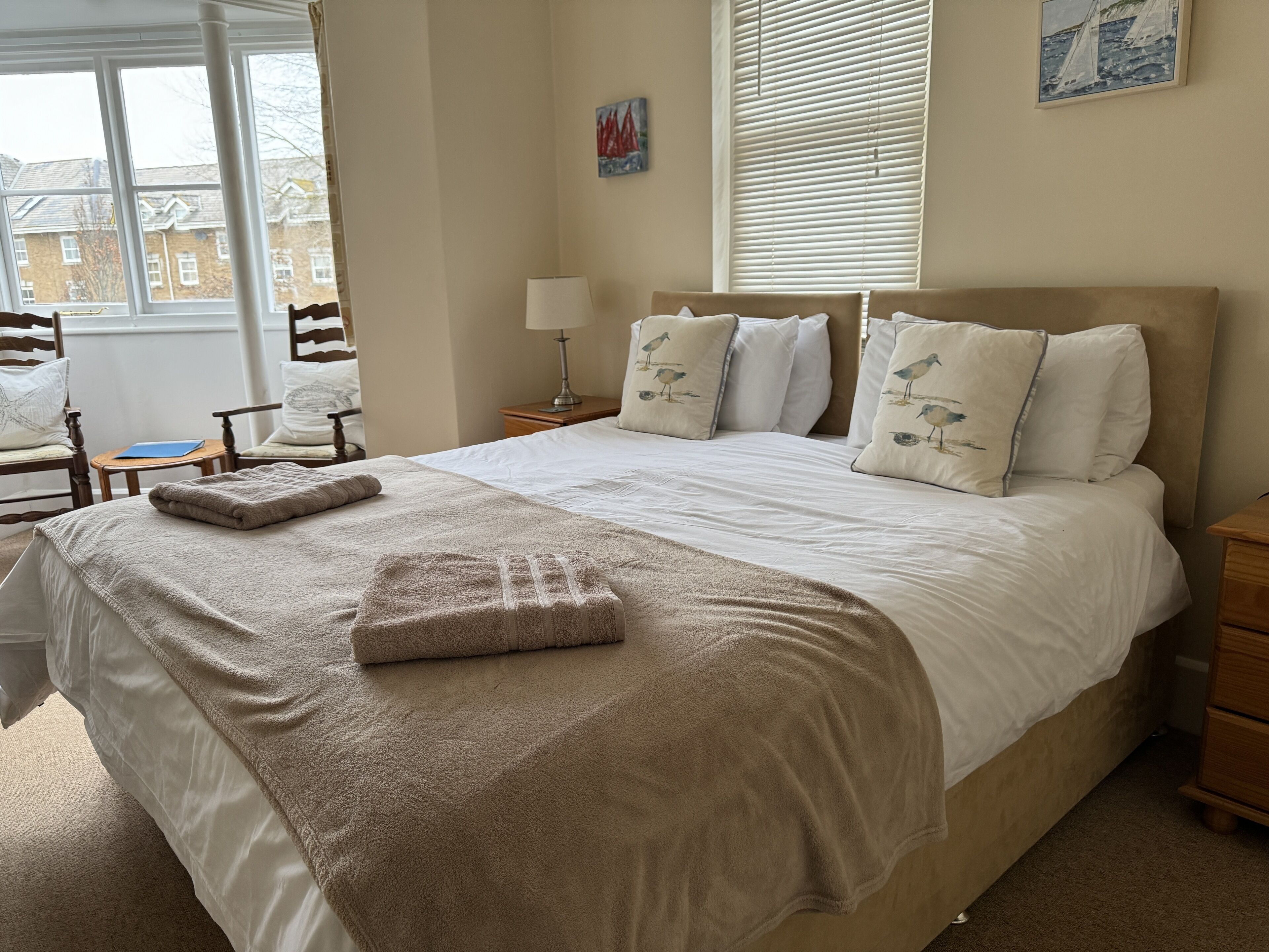 A neatly made bed with a beige blanket and pillows, a bedside lamp, and a window with blinds.