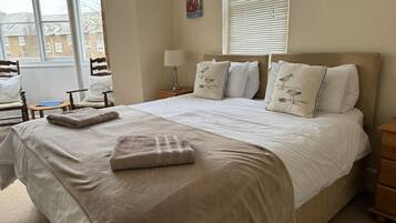 A neatly made bed with a beige blanket and pillows, a bedside lamp, and a window with blinds.