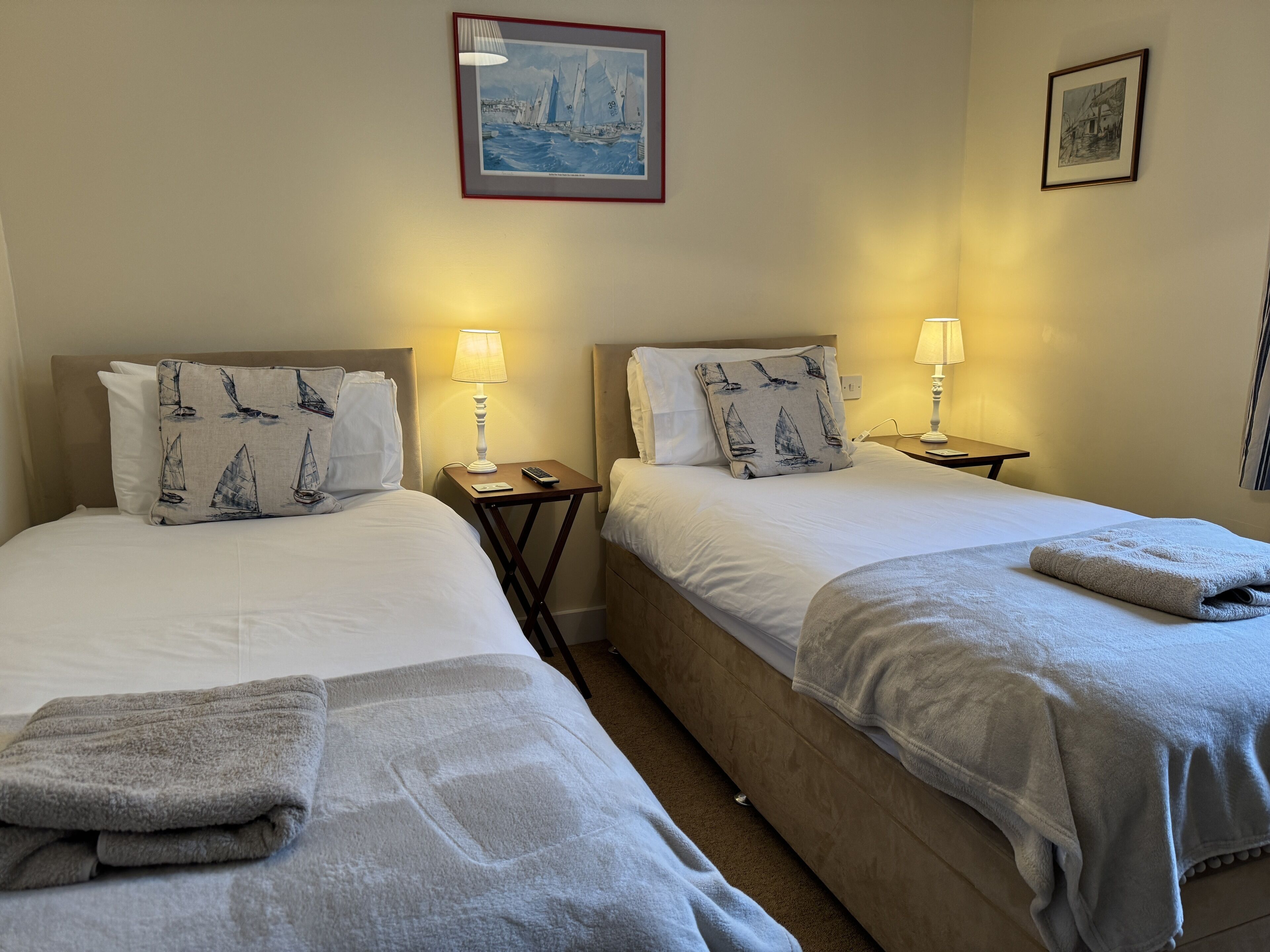 A hotel room with two beds, each with a pillow featuring sailboats, a bedside table with a lamp, and a painting on the wall.