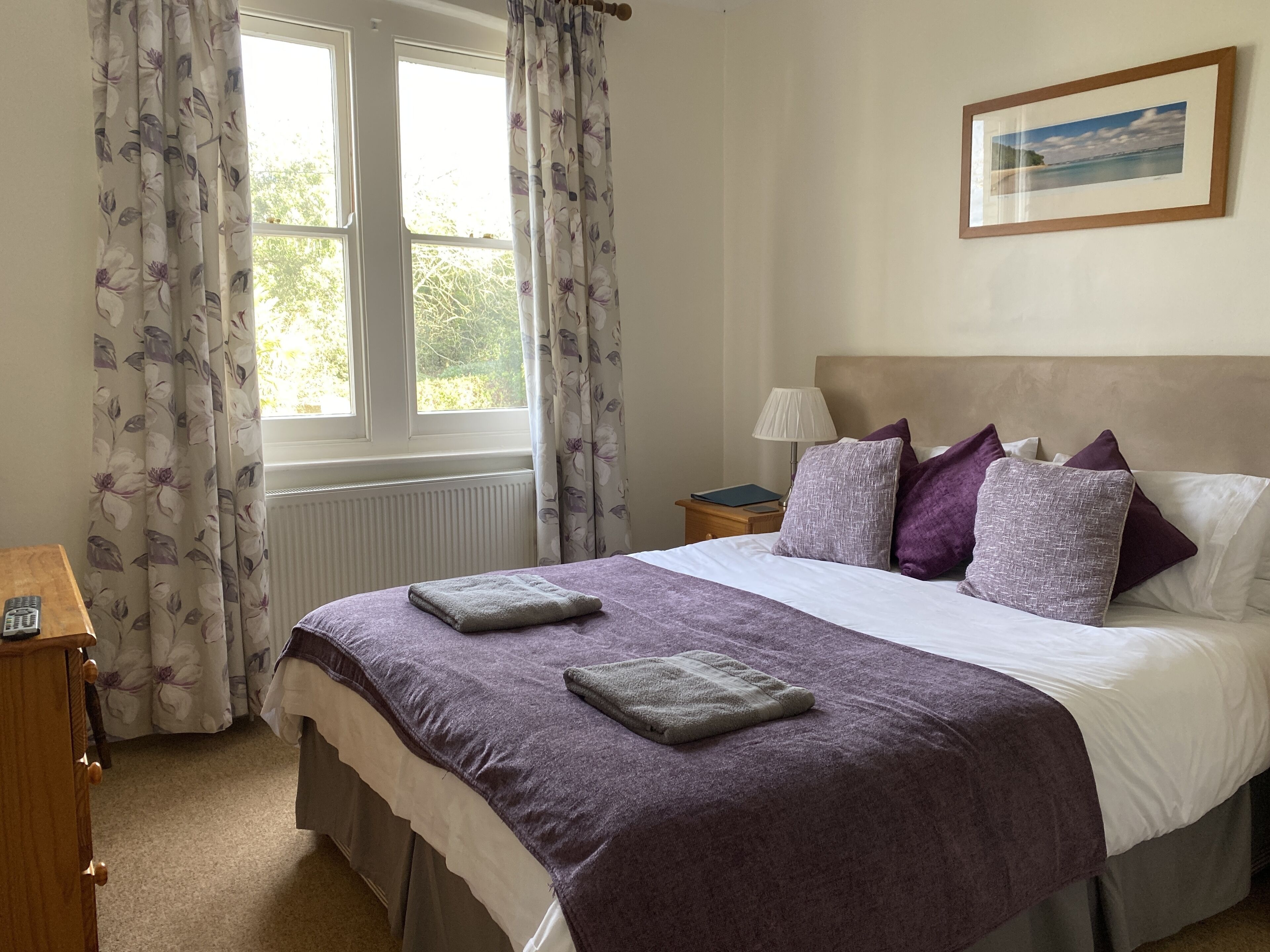 A neatly arranged bedroom with a bed, pillows, a bedside lamp, a framed picture on the wall, and a window with curtains.