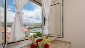 A room with a large window, white curtains, and a vase with red and yellow flowers on a wooden table.