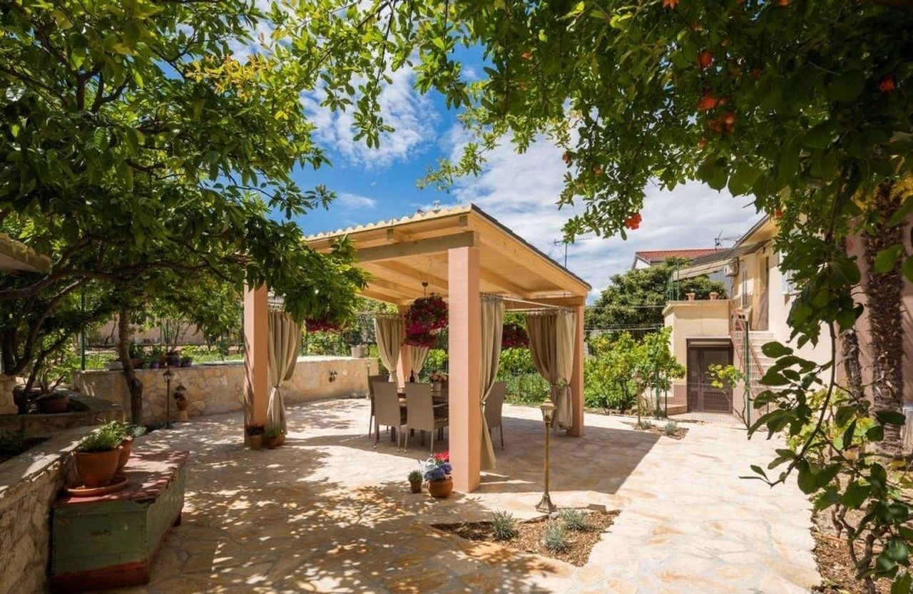 A covered outdoor patio with a table and chairs, surrounded by potted plants and a stone floor.